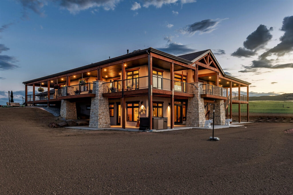 Beautiful evening shot of a huge two-story mansion with a wraparound porch with massive stone pillars supporting. In the background, the Colorado sunset is beginning to occur across the front range.